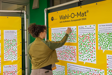 Person mit braunen Locken klebt einen Klebepunkt auf eine These auf der gelben Wahl-O-Mat Plane zur Europawahl 2024