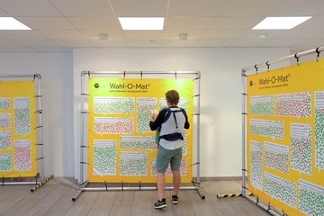 Person mit Kleinkind an Brust geschnall klebt Punkte auf Wahl-O-Mat Plane mit Thesen zur Europawahl 2024