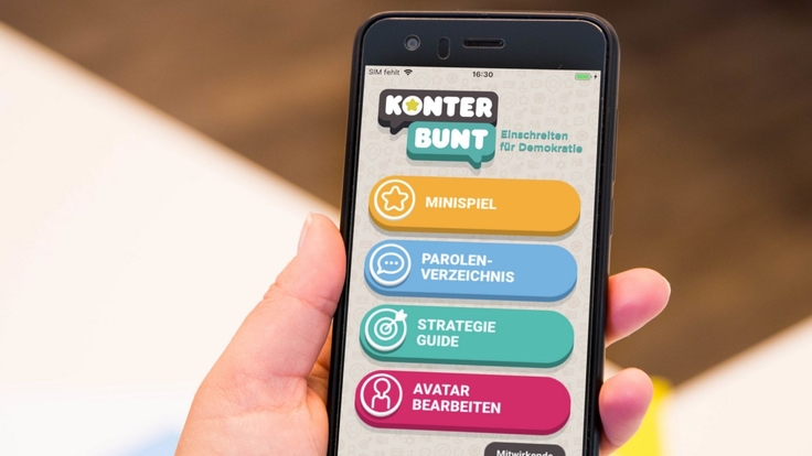 Ein Smartphone in einer Hand, auf dem Bildschirm ist zu lesen "KonterBUNT" Einschreiten für Demokratie. Im Menü stehen zur Auswahl "Mnispiel", "Parolenverzeichnis", "Strategie Guide" und "Avatar bearbeiten&a