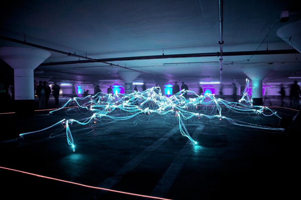 Ein dunkles Parkhaus, erleuchtet von Linien aus Licht, die ein Netzwerk bilden dazu laufen Menschen im Hintergrund, Symbolbild Digitalisierung