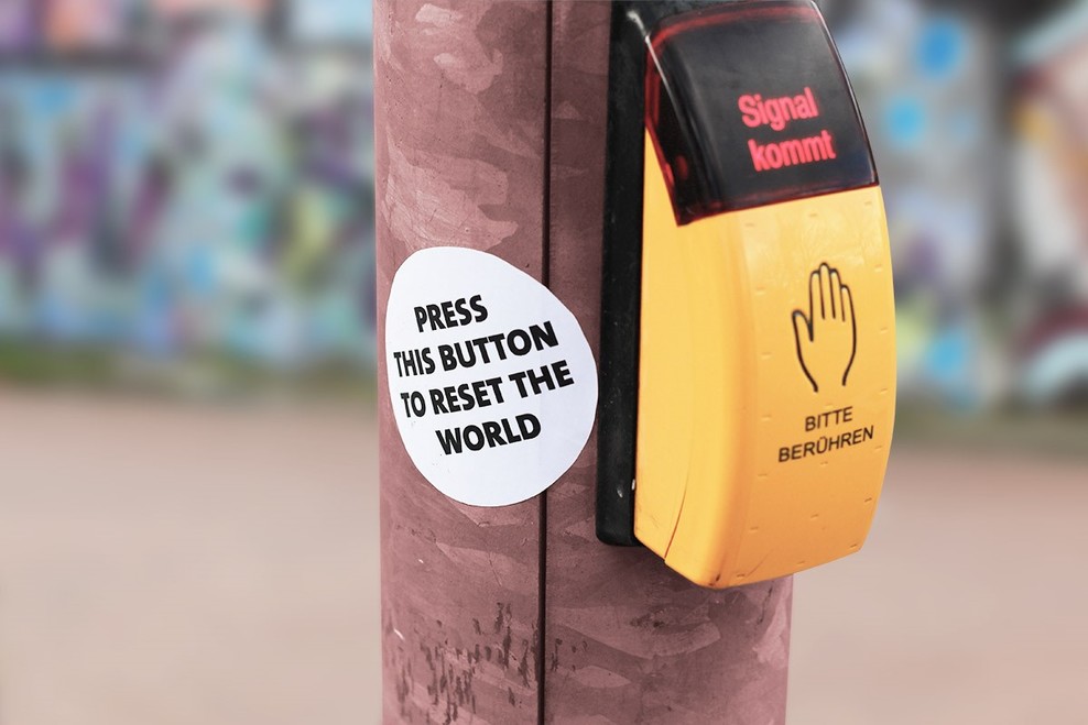 Zu sehen ist ein gelber Knopf, mit dem Fußgänger_innen Ampeln aktivieren können. Daneben ein Aufkleber mit der Aufschrift "Press this button to reset the world"
