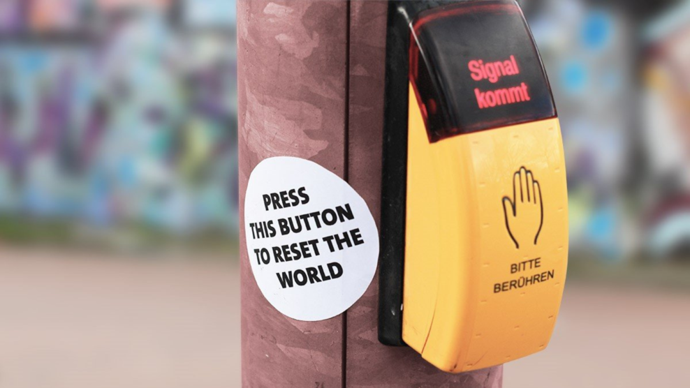 Zu sehen ist ein gelber Knopf, mit dem Fußgänger_innen Ampeln aktivieren können. Daneben ein Aufkleber mit der Aufschrift "Press this button to reset the world"