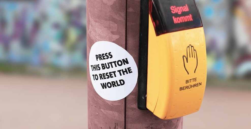 Zu sehen ist ein gelber Knopf, mit dem Fußgänger_innen Ampeln aktivieren können. Daneben ein Aufkleber mit der Aufschrift "Press this button to reset the world"