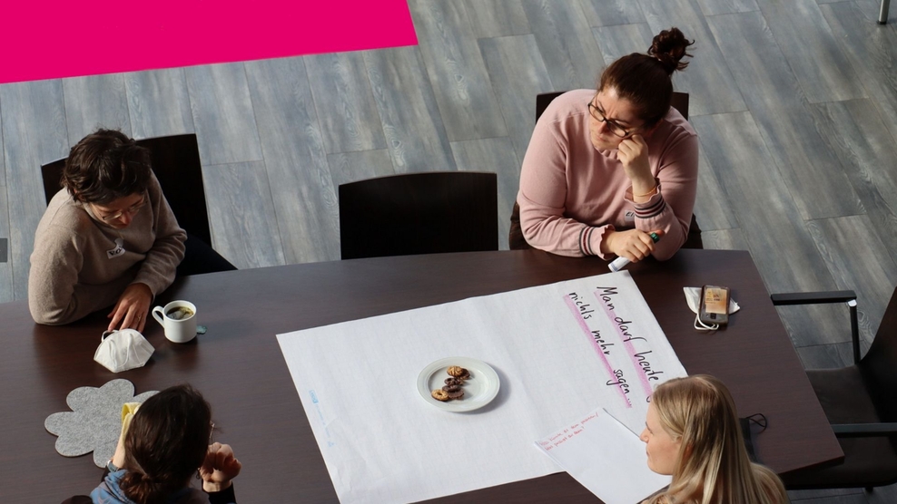 Eine Gruppe von Personen sitzt um einen Tisch, auf dem ein großes Blatt Papier liegt.